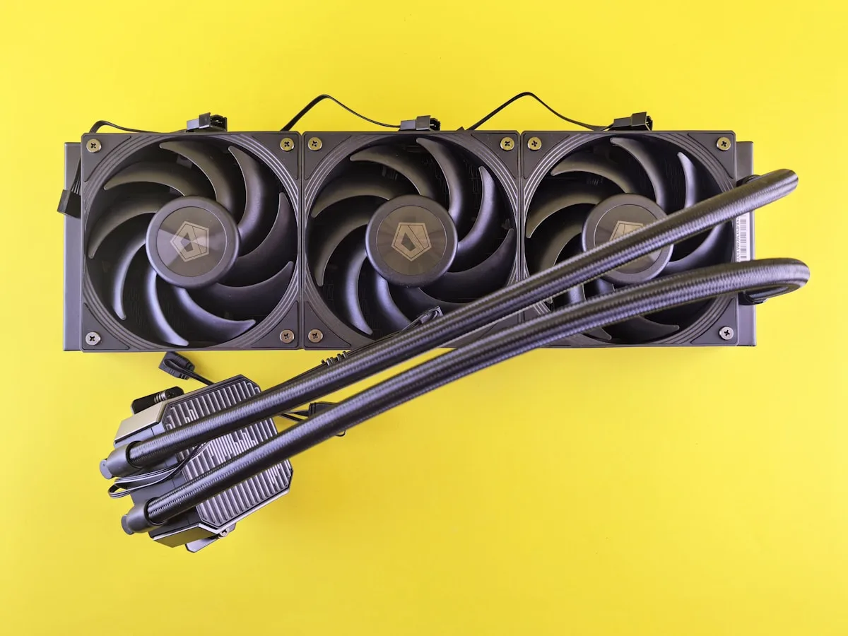 Radiador de water cooler de 360 mm com três ventoinhas pretas ao lado de um cooler a ar dual-tower com dissipador de alumínio, lado a lado sobre fundo amarelo