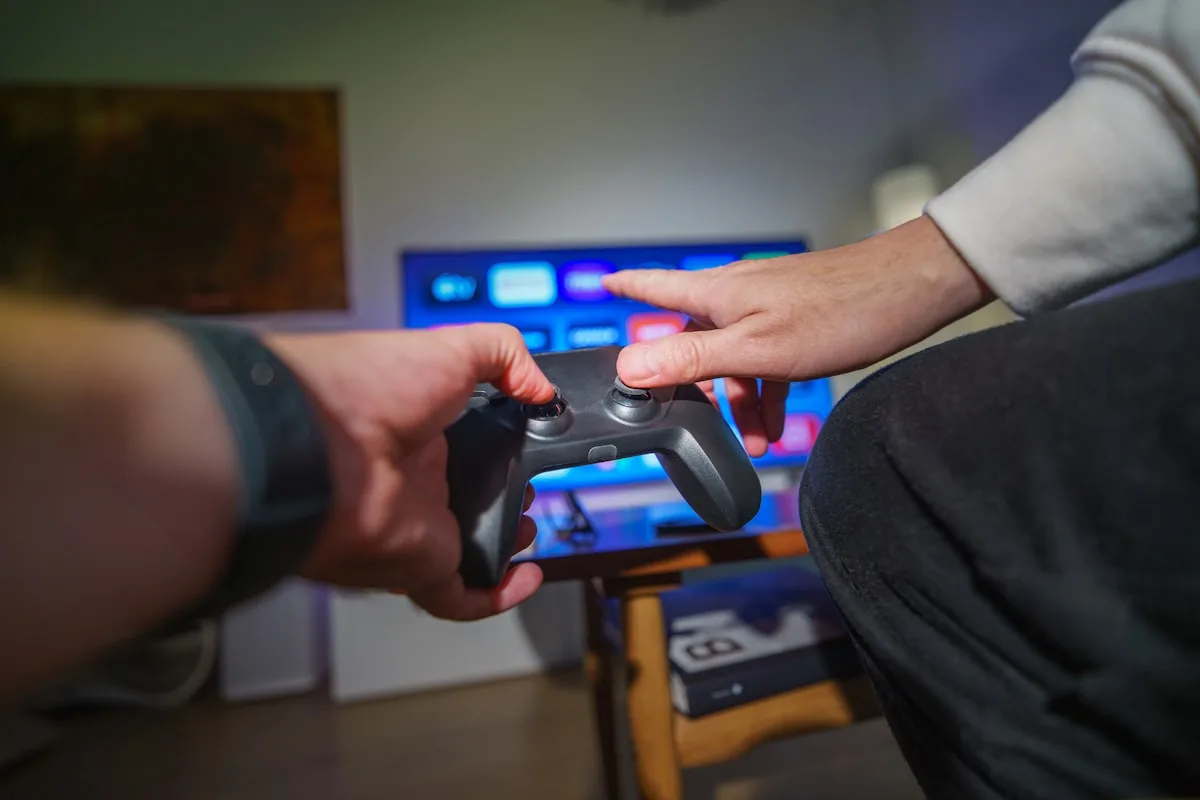 Duas mãos passando um controle de video game em frente à TV ligada em ambiente doméstico