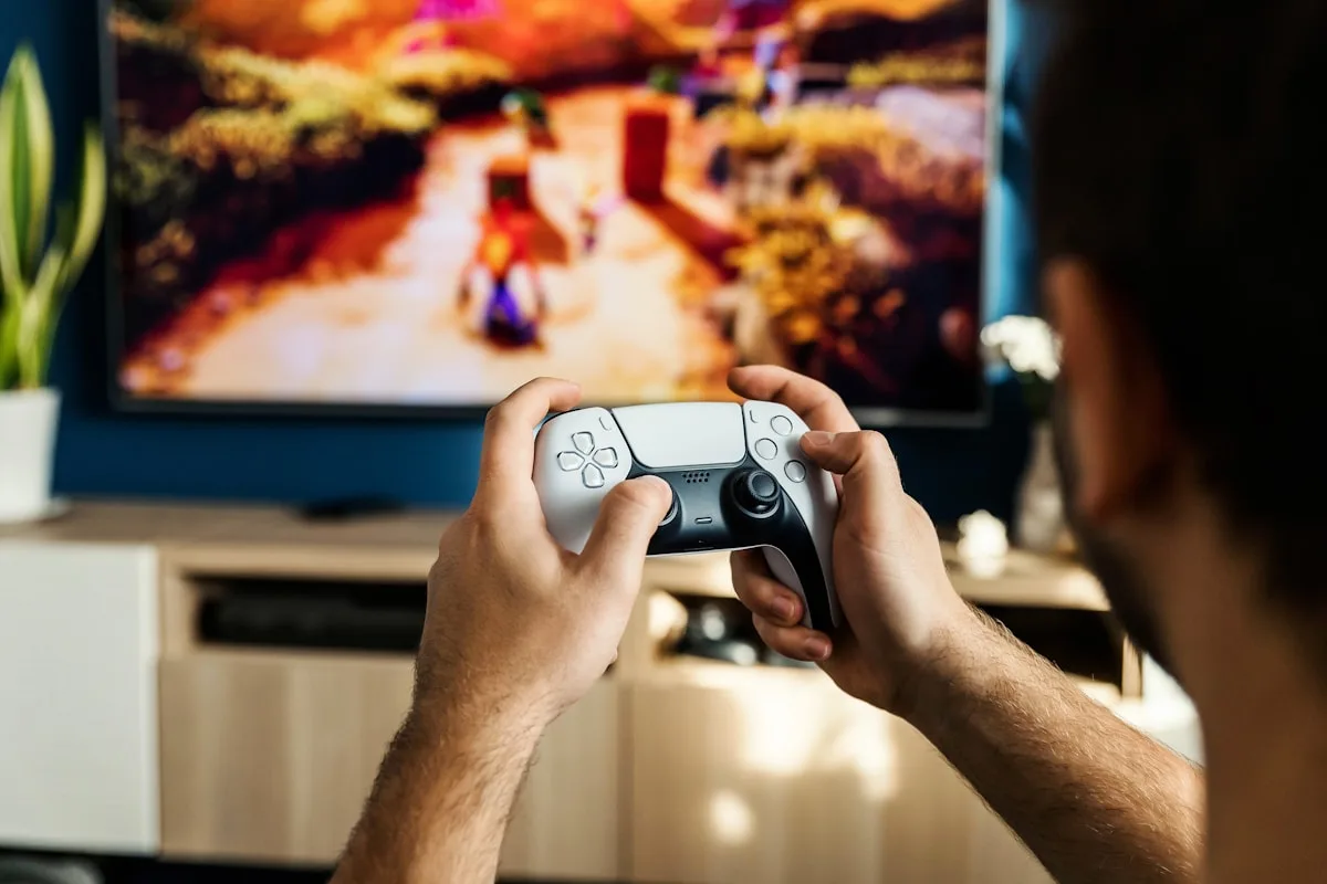 Jogador segurando controle sem fio branco em frente à TV com cena de jogo desfocada ao fundo