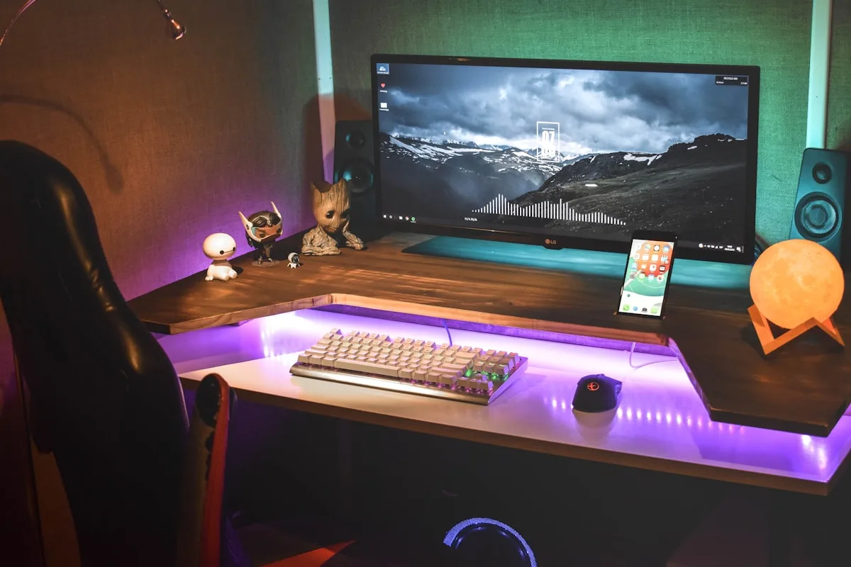 Fita de LED cobrindo vários pontos do setup Mesa em L com fita de LED roxa embaixo iluminando o chão e luz teal atrás do monitor