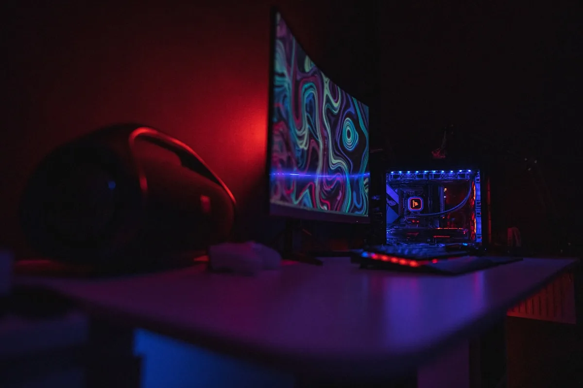 Mesa gamer escura com monitor exibindo cores em movimento e luz ambiente vermelha na parede