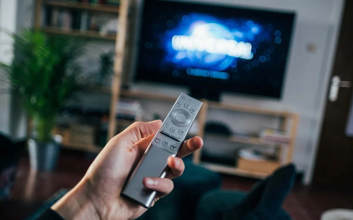 Controle de smart TV em uso Mão segura um controle de smart TV com uma tela azul desfocada ao fundo em uma sala de estar