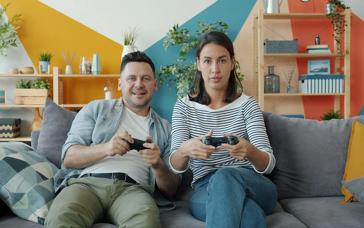 Um mundo aberto maior à espera do multiplayer Duas pessoas jogam vídeogame sentadas no sofá, controles em mãos e parede colorida ao fundo