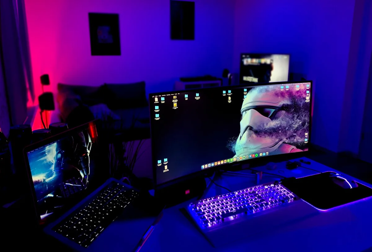 Tipos de iluminação para setup gamer Setup gamer com dois monitores e luz ambiente azul e rosa refletindo nas paredes do quarto