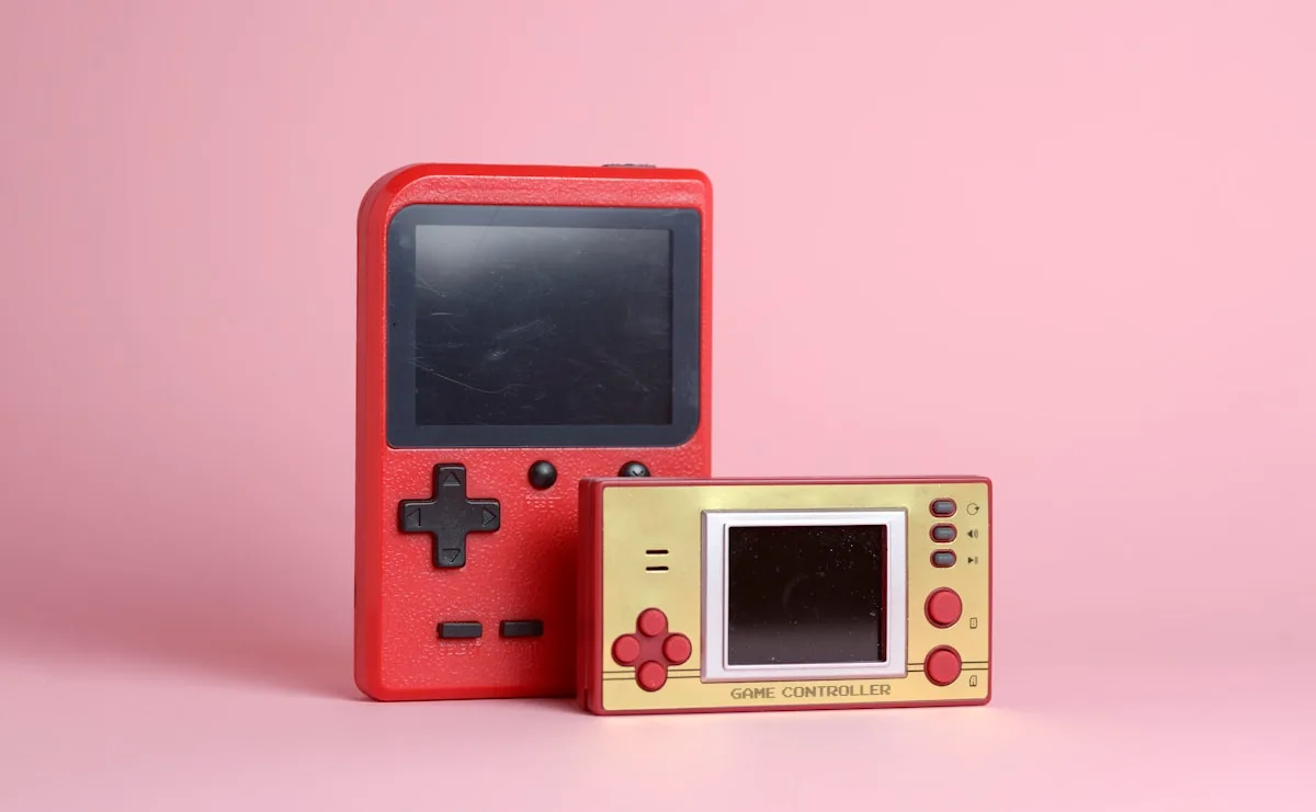 Dois handhelds retrô compactos, vermelho e dourado, lado a lado sobre fundo rosa