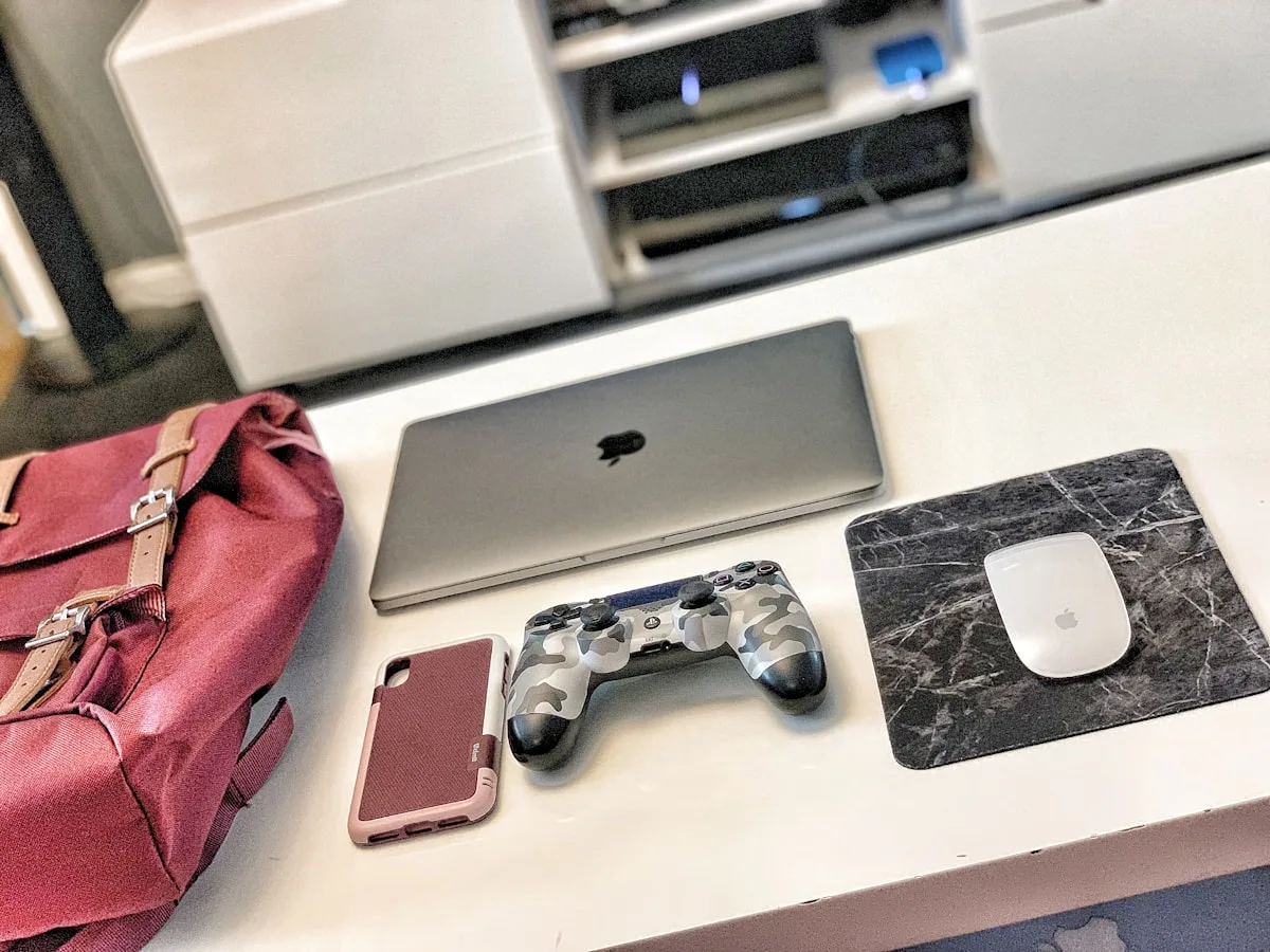 Mochila gamer vinho ao lado de notebook, controle de PS4 e mouse sobre mesa branca