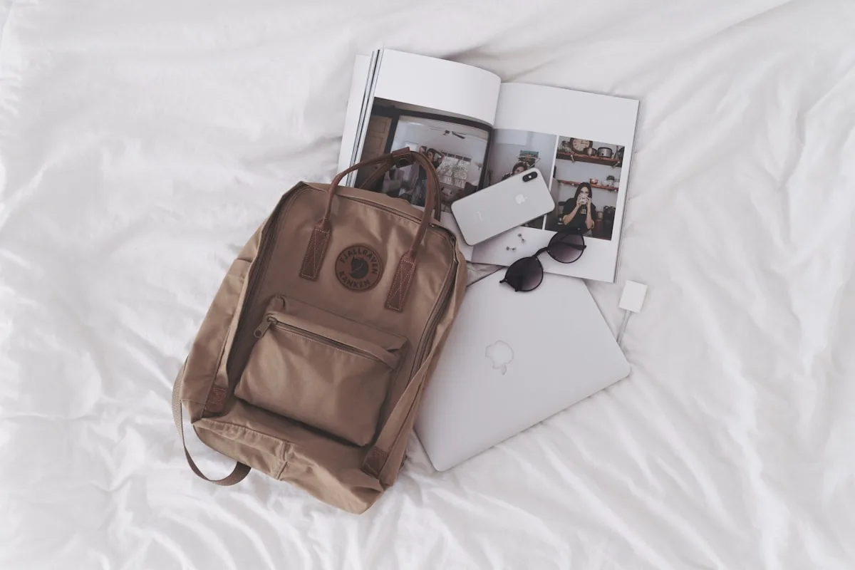 Mochila compacta bege sobre a cama ao lado de notebook, celular e óculos de sol