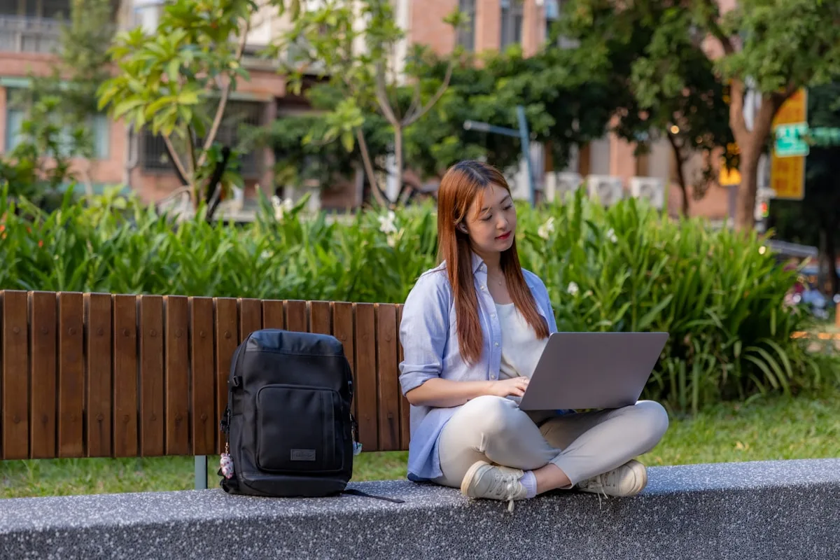 Mochila preta apoiada em banco de parque enquanto usuária trabalha no notebook ao ar livre