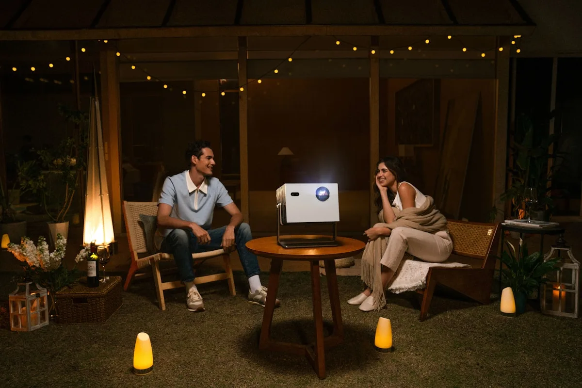 Sessão de jogo com projetor portátil ao ar livre Casal sentado ao redor de mesa com projetor portátil branco ligado em jardim durante a noite
