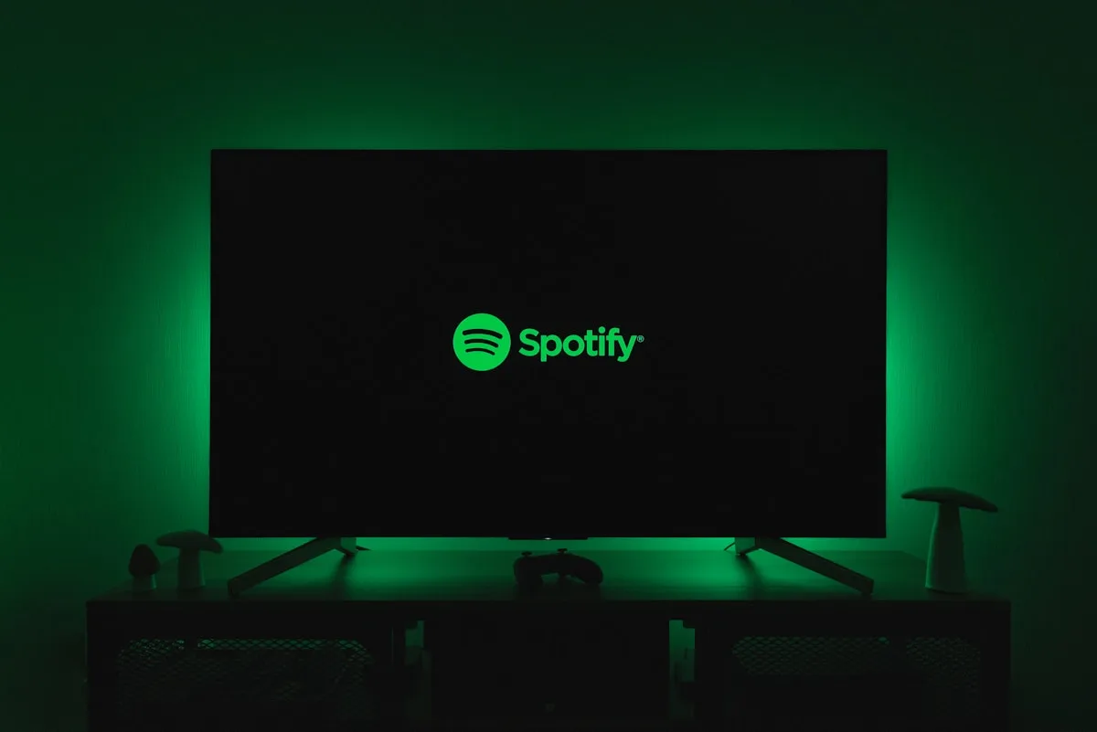 TV com luz de fundo verde, tela do Spotify e controle de videogame apoiado no rack