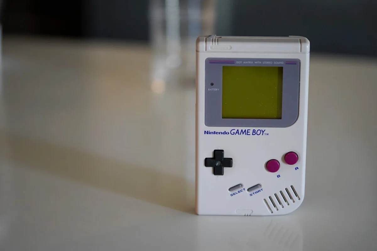 Game Boy original branco em foco sobre mesa clara, com tela retroiluminada em verde
