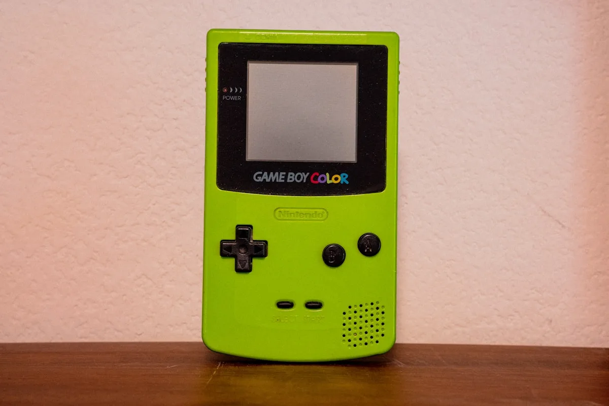 Game Boy Color verde-limão apoiado em parede clara, com D-pad e botões de ação visíveis