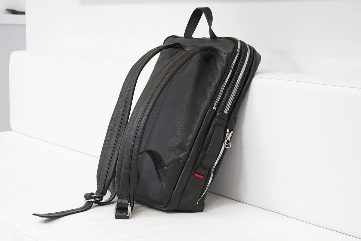 Mochila preta de couro apoiada em bloco branco mostrando zíper duplo reforçado e alças traseiras