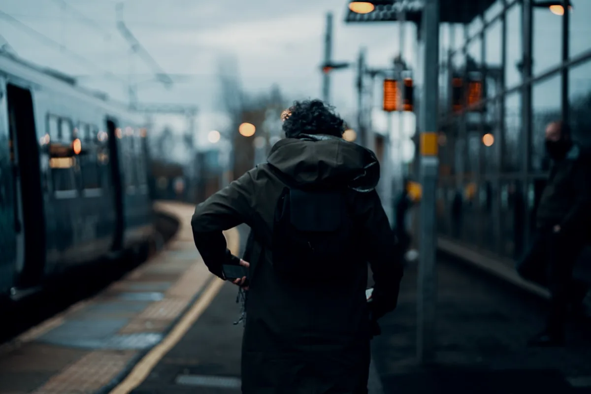 Pessoa de costas em plataforma de trem ao anoitecer com mochila preta e casaco escuro, aguardando embarque