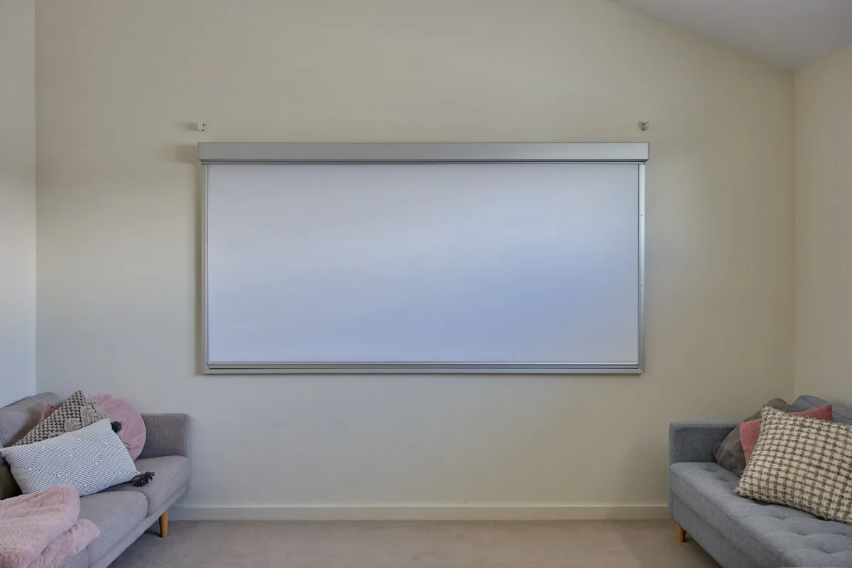 Tela de projeção instalada em sala de estar Tela de projeção retrátil branca instalada na parede de uma sala de estar, acima de um sofá cinza com almofadas