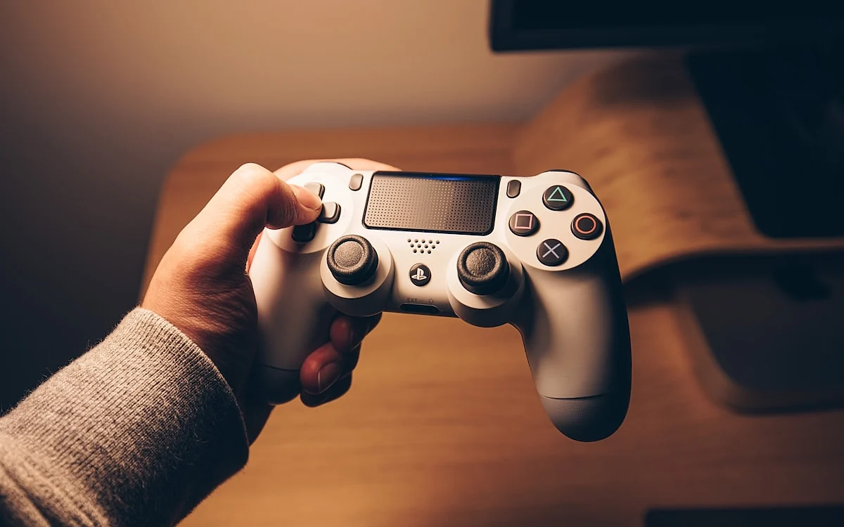 TV e console formam o mesmo investimento Mão segurando um controle de videogame branco perto de uma mesa, com uma tela desfocada ao fundo