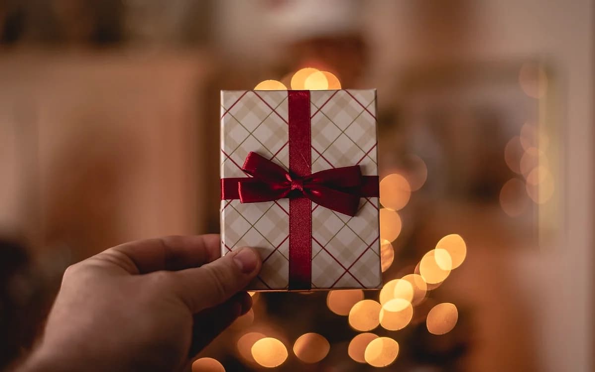Gift Card PSN para GTA 6 Mão segurando um presente embrulhado com laço vermelho e luzes desfocadas ao fundo