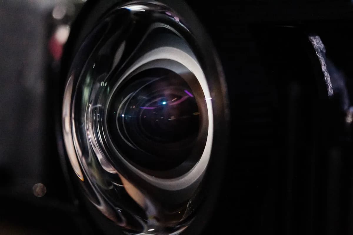 4K Nativo ou Pixel Shift: Qual Projetor Comprar em 2026 Lente de projetor em close, com reflexos de luz nos elementos de vidro sobre fundo escuro