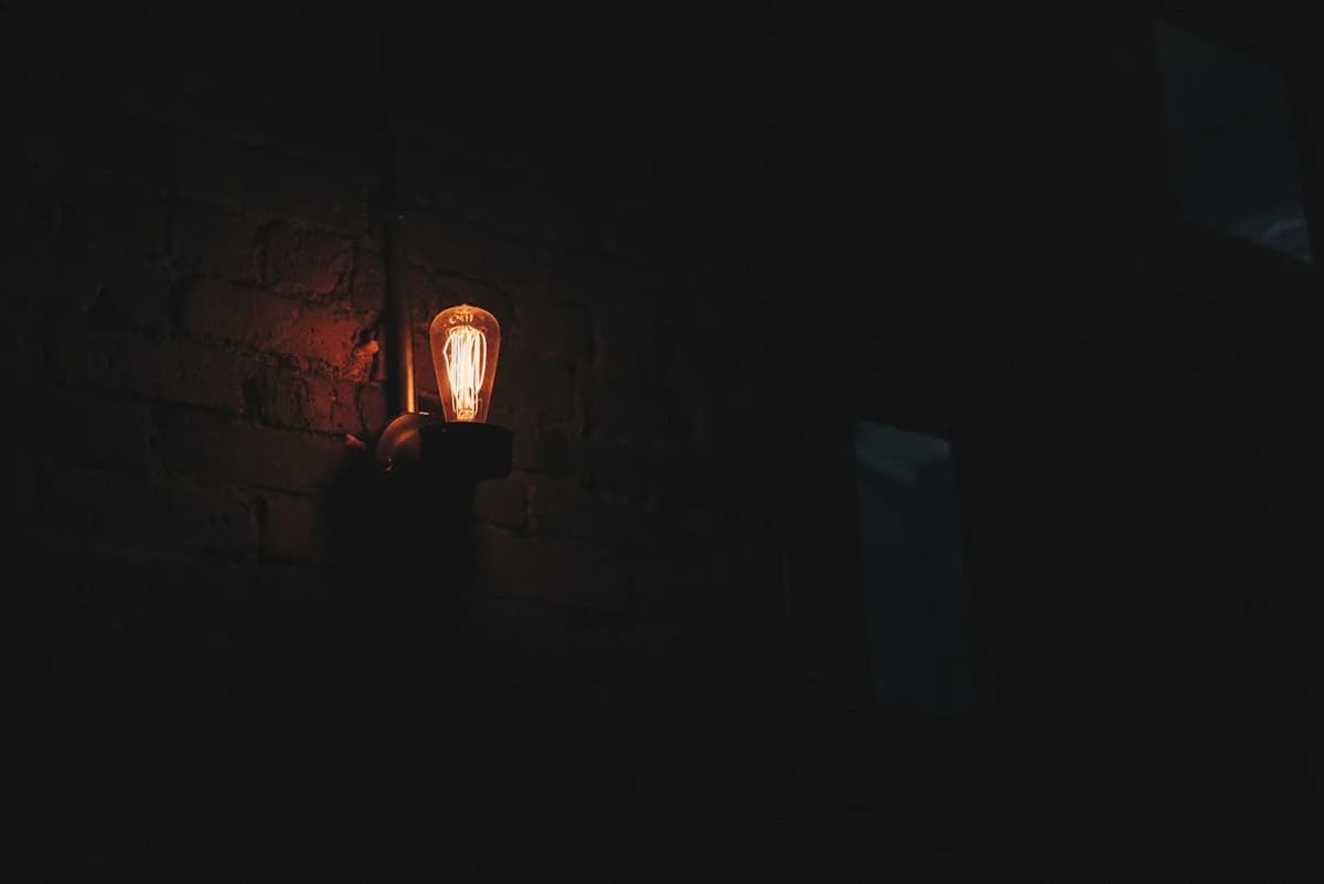 Projetor para Jogos Vale a Pena em 2026 Lâmpada incandescente acesa sobre parede de tijolos escura, único ponto de luz em ambiente sem iluminação