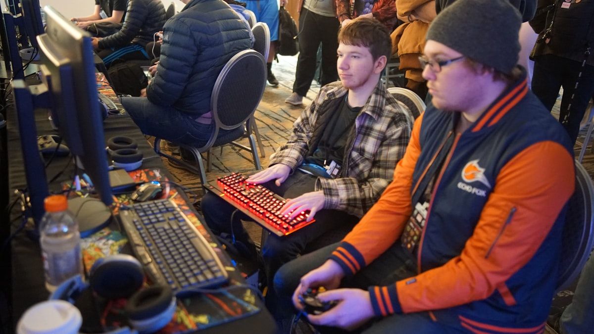 Jogadores se divertindo com controle e teclado em 2026 Homem jogando videogame com controle ao lado de outro usando teclado no computador